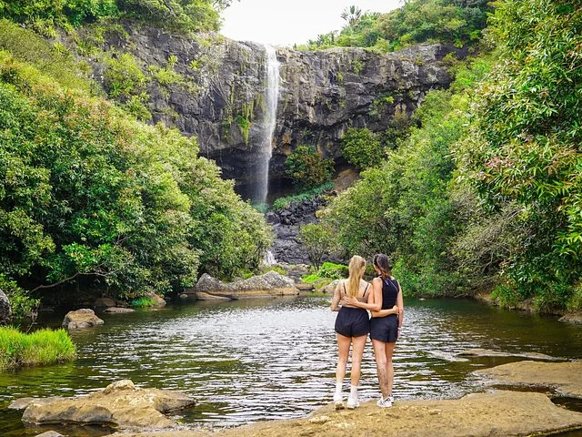 Two women standing on rocks near a pool facing a tall waterfall